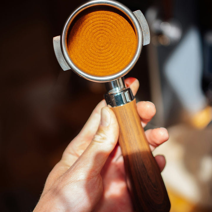 A barista's hand holding an espresso portafilter loaded with freshly ground coffee, about to dose and tamp before pulling a shot