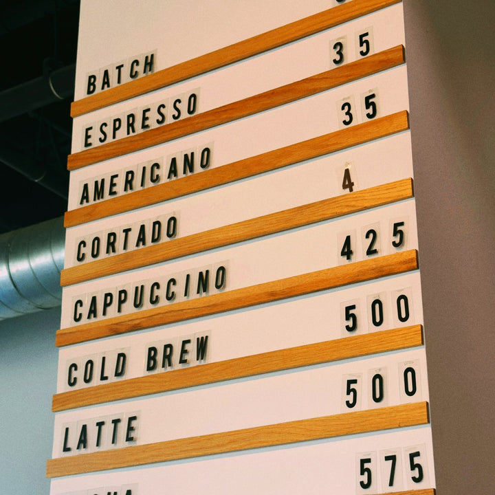 A cafe menu board of wooden rails and black plastic letters listing each drink as its own priced line - Inheritance Coffee, Pensacola, FL