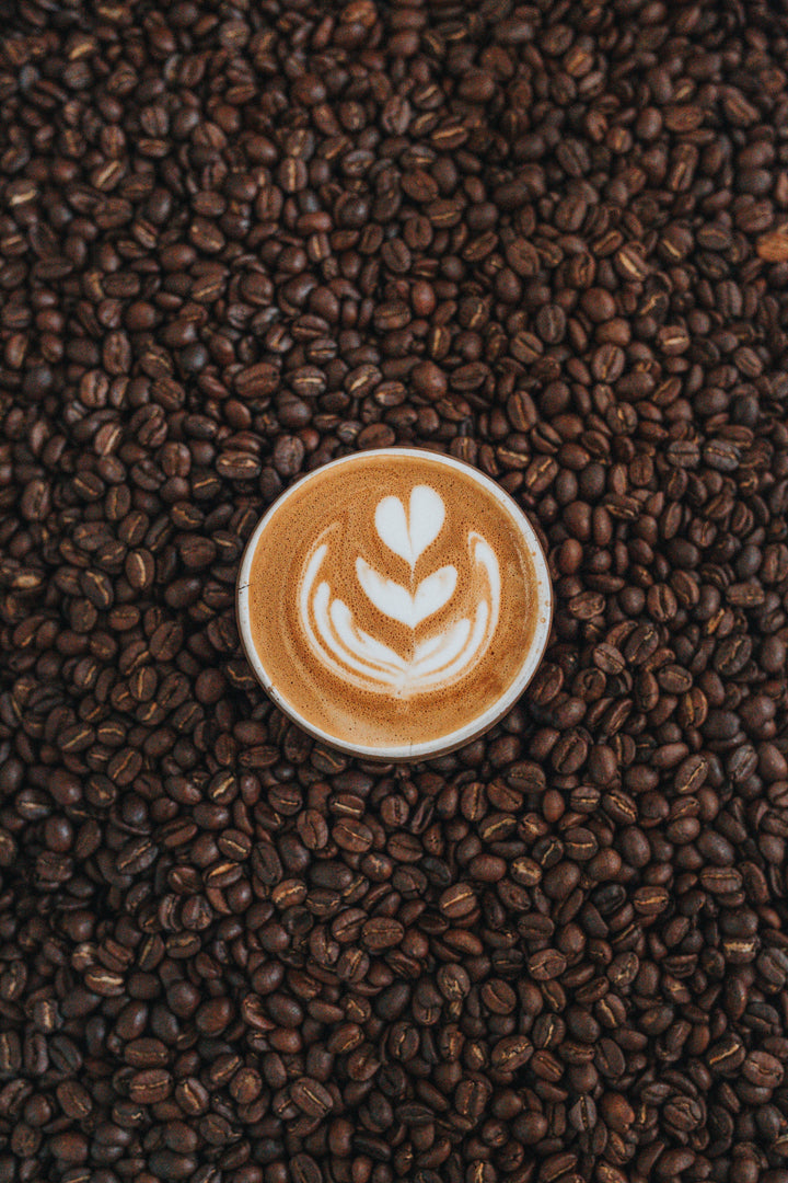 Latte art in a cup surrounded by roasted coffee beans - home brewing guide from Inheritance Coffee, Pensacola, FL
