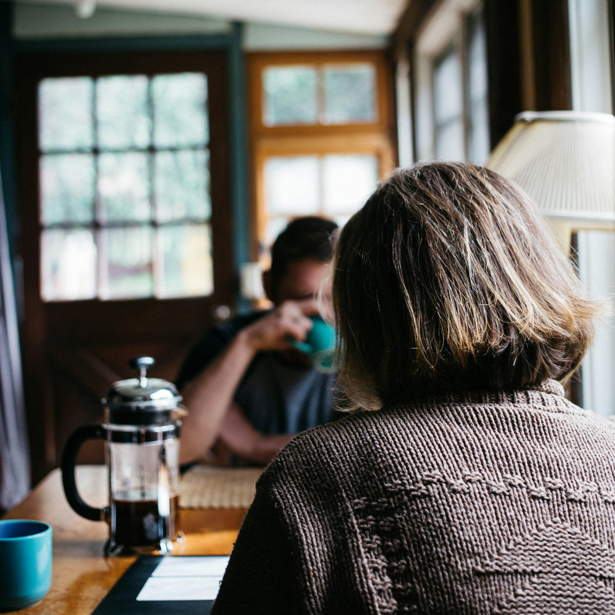 A French press and two mugs of coffee on a cafe table between two people talking