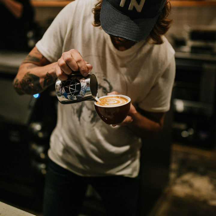 Barista pouring steamed milk from a metal pitcher into a cup of espresso on a cafe bar