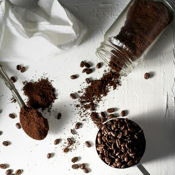 Whole roasted coffee beans and freshly ground coffee laid out beside a cup of black coffee on a white surface