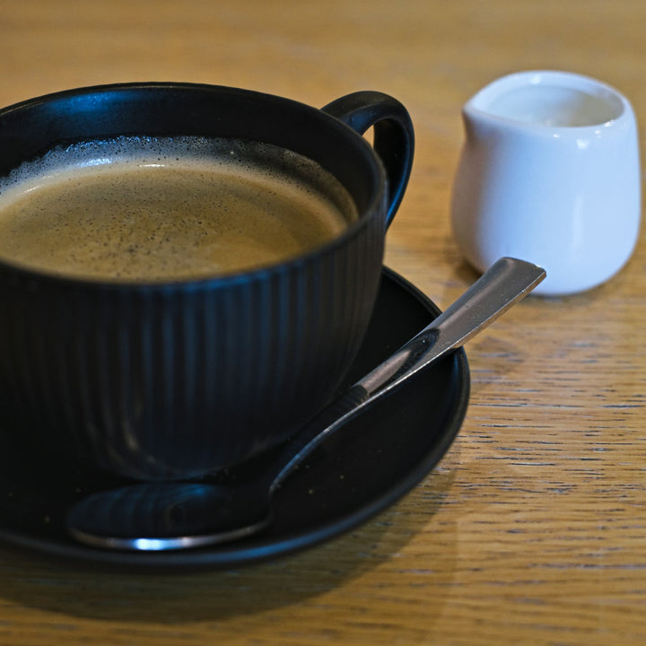 A black mug of coffee on a saucer with a biscotti and creamer