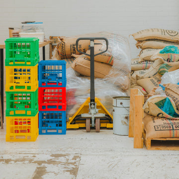 Sacks of green coffee stacked on pallets in a warehouse with a pallet jack alongside