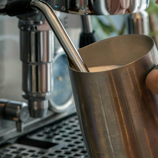 A barista's hand wrapped around a stainless steel milk pitcher while steaming milk on an espresso machine