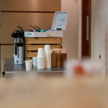 A stainless steel coffee urn and stacked paper cups set out as a refreshment station for a conference break