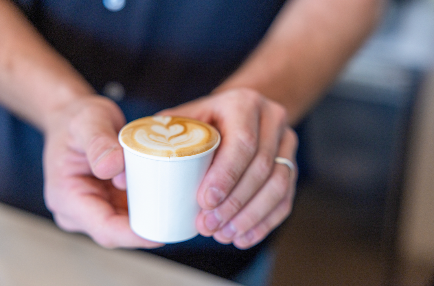 Person holding a white cup with a cappuccino in a blurred indoor setting