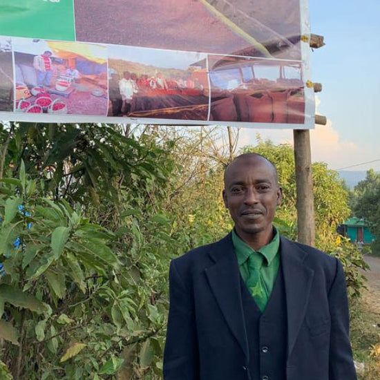 Man standing in front of a wooden structure with a signboard displaying images, with a vehicle and motorcycle in the background.
