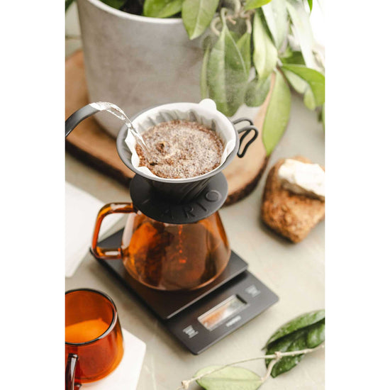 Coffee brewing setup with a pour-over coffee maker on a digital scale, surrounded by a plant and toast.