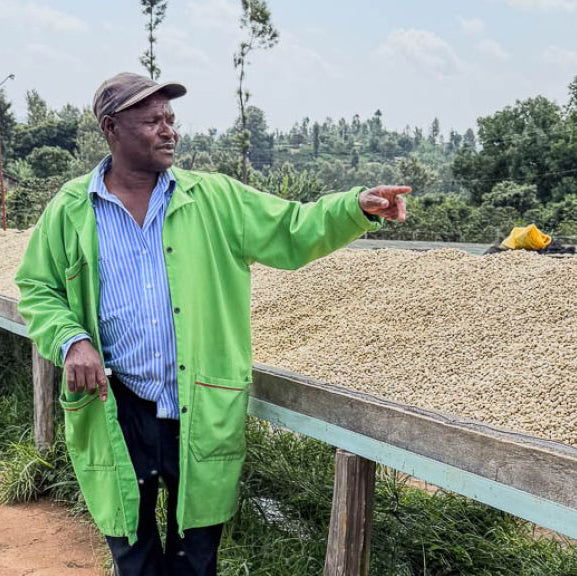 Man in a green jacket standing next to a table with drying coffee beans outdoors.